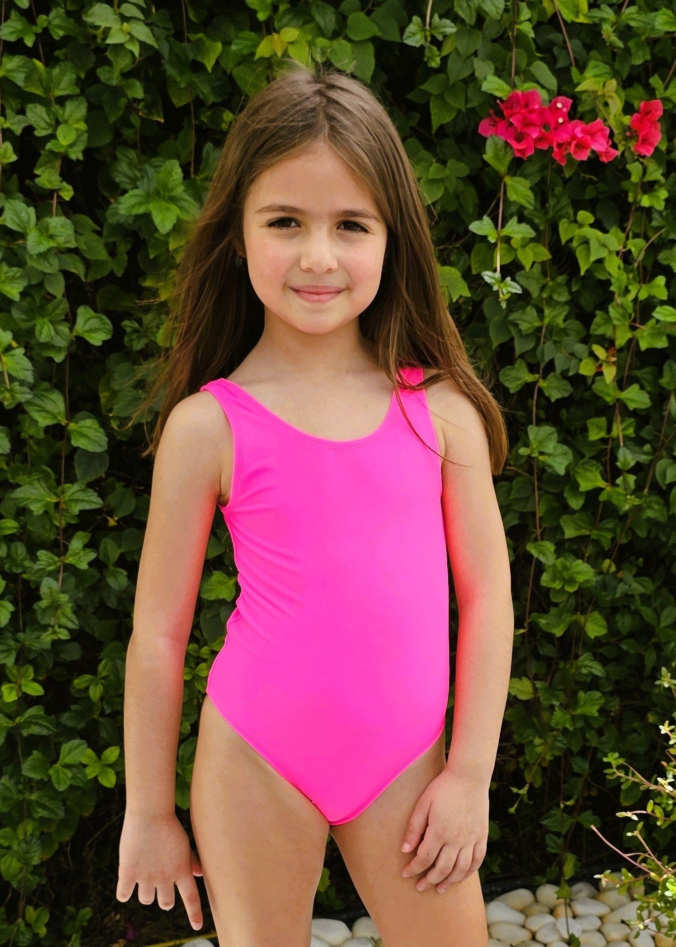 Young girl in a pink swimsuit standing outdoors with greenery in the background